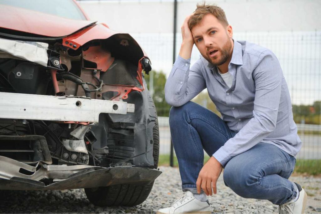 Man crouching beside a car with visible front-end damage, illustrating post-accident confusion and the need to document vehicle damage after a rideshare collision.