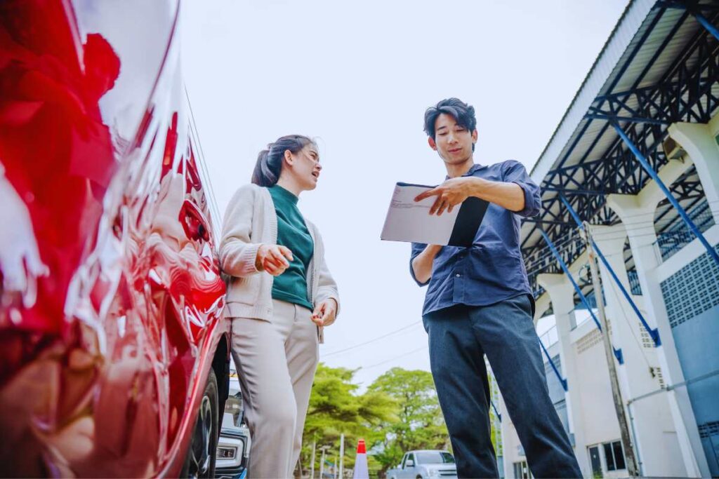 Driver and passenger reviewing documents beside a dented red car after a rideshare accident, showing the importance of collecting information at the scene.