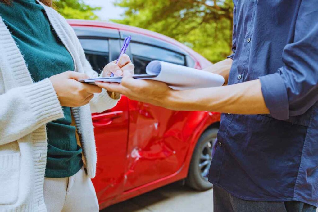 Person writing on a clipboard while another person holds paperwork near a damaged red car, representing the exchange of insurance or accident information after a collision.