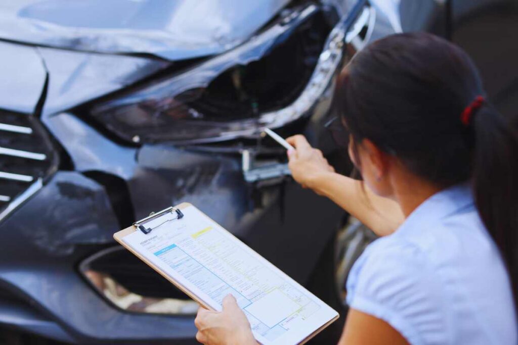 Person holding a clipboard while inspecting front-end vehicle damage, illustrating how insurance adjusters document property damage after a car accident.