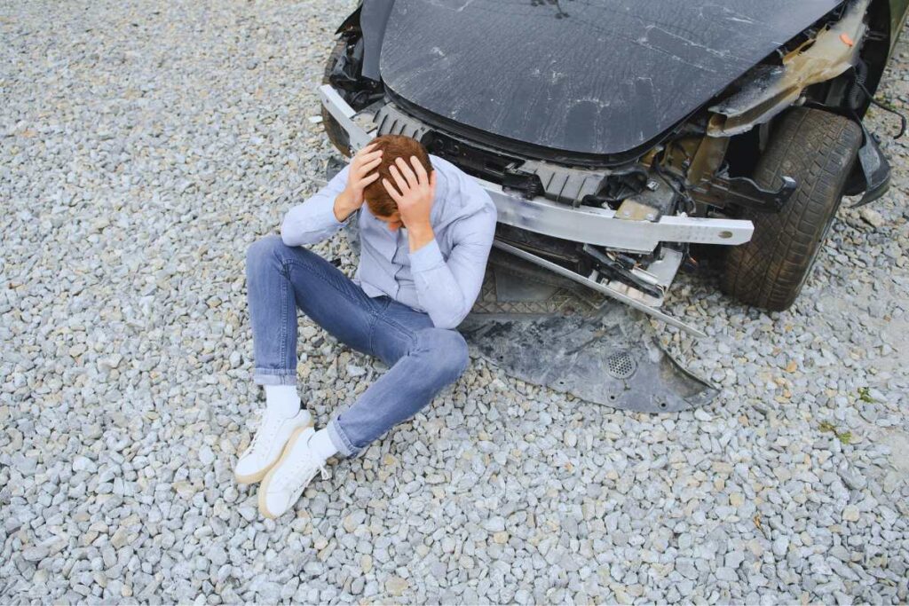 Man sitting on gravel holding his head in pain beside a badly damaged vehicle after a rideshare accident, illustrating the immediate confusion and injury risks in San Diego crashes.