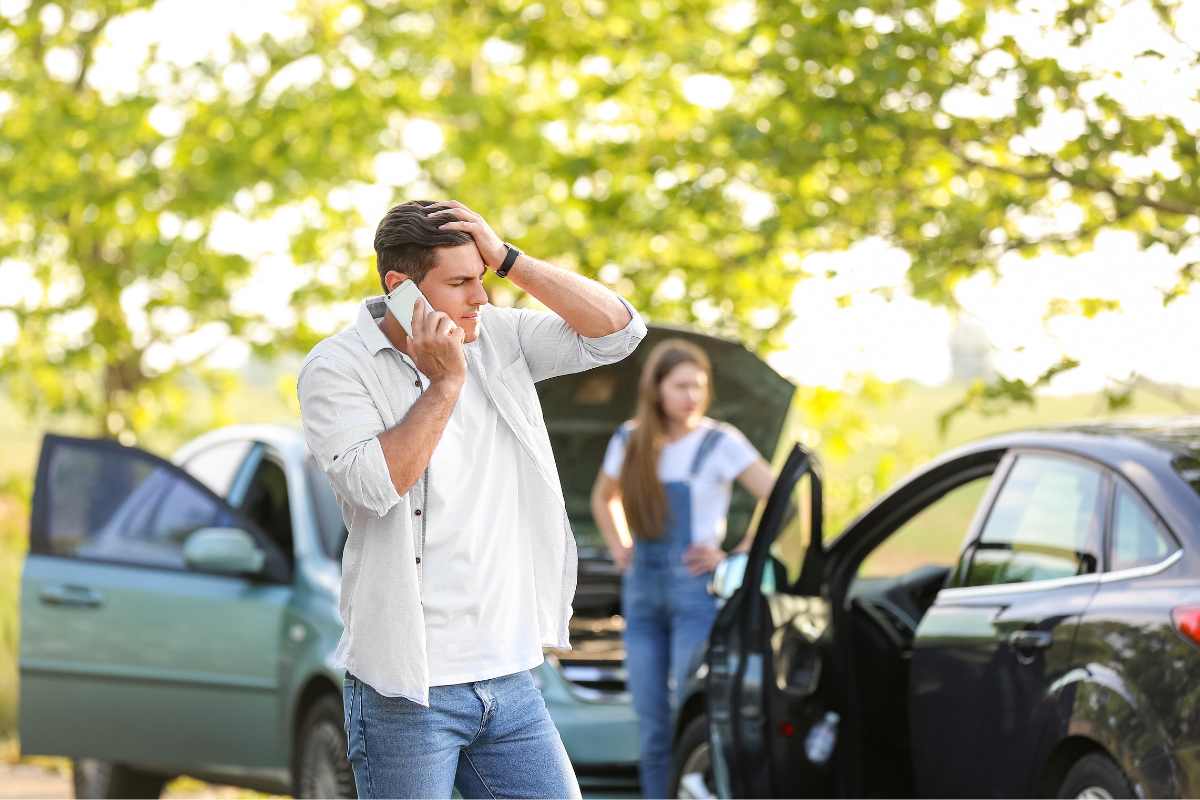 A distressed man making a phone call after a car accident, with damaged vehicles and a concerned passenger in the background, symbolizing the stress of post-crash medical bills in San Diego.