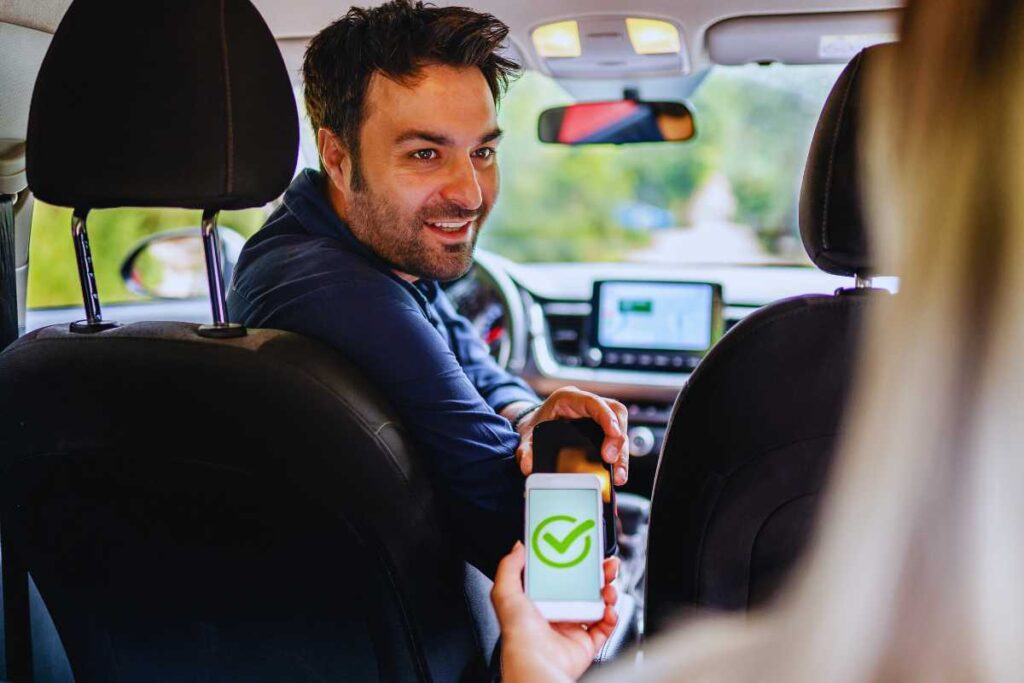 Uber driver seated in a car showing a smartphone with a confirmed ride status to a passenger, illustrating how trip status affects rideshare insurance coverage after a crash.